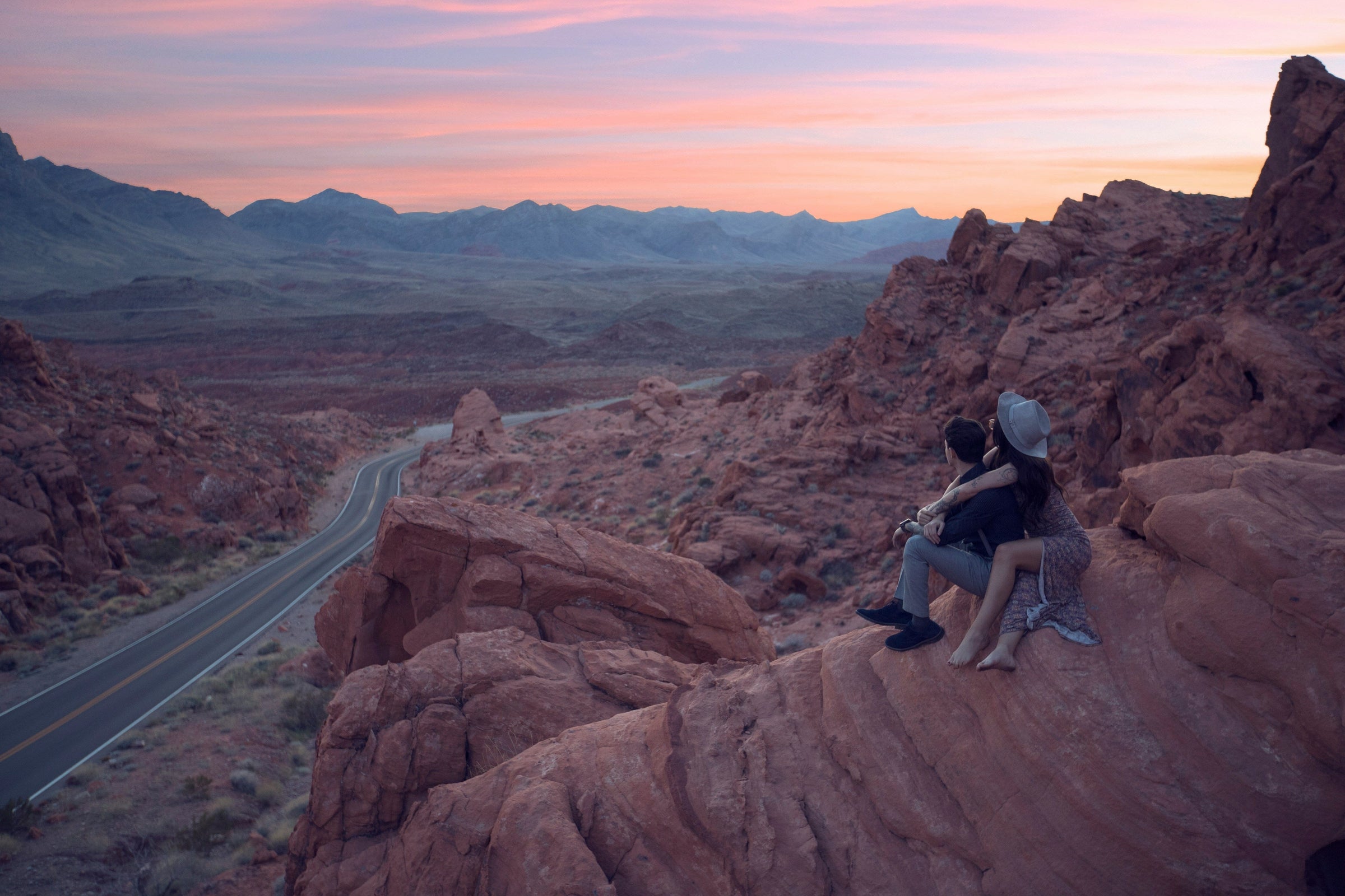 Deux voyageurs contemplant un paysage au soleil couchant avec une route qui s'étire vers l'horizon, illustrant l'évasion et la promesse d'aventure d'une carte de voyage Cartovia.